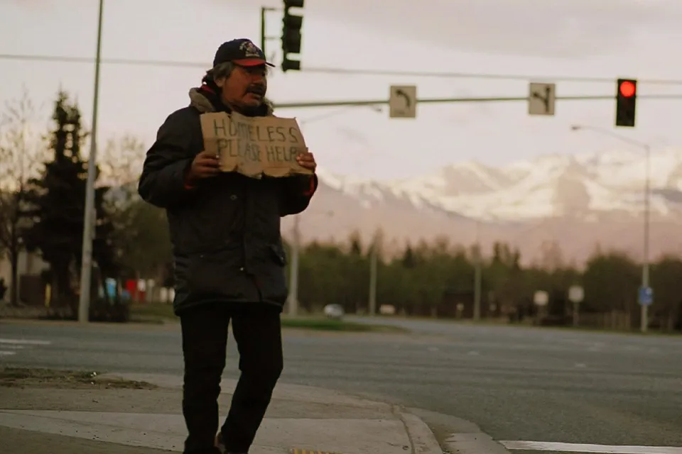 Unhoused man holds up cardboard sign on street corner in Anchorage, Alaska with snowy mountains in background.